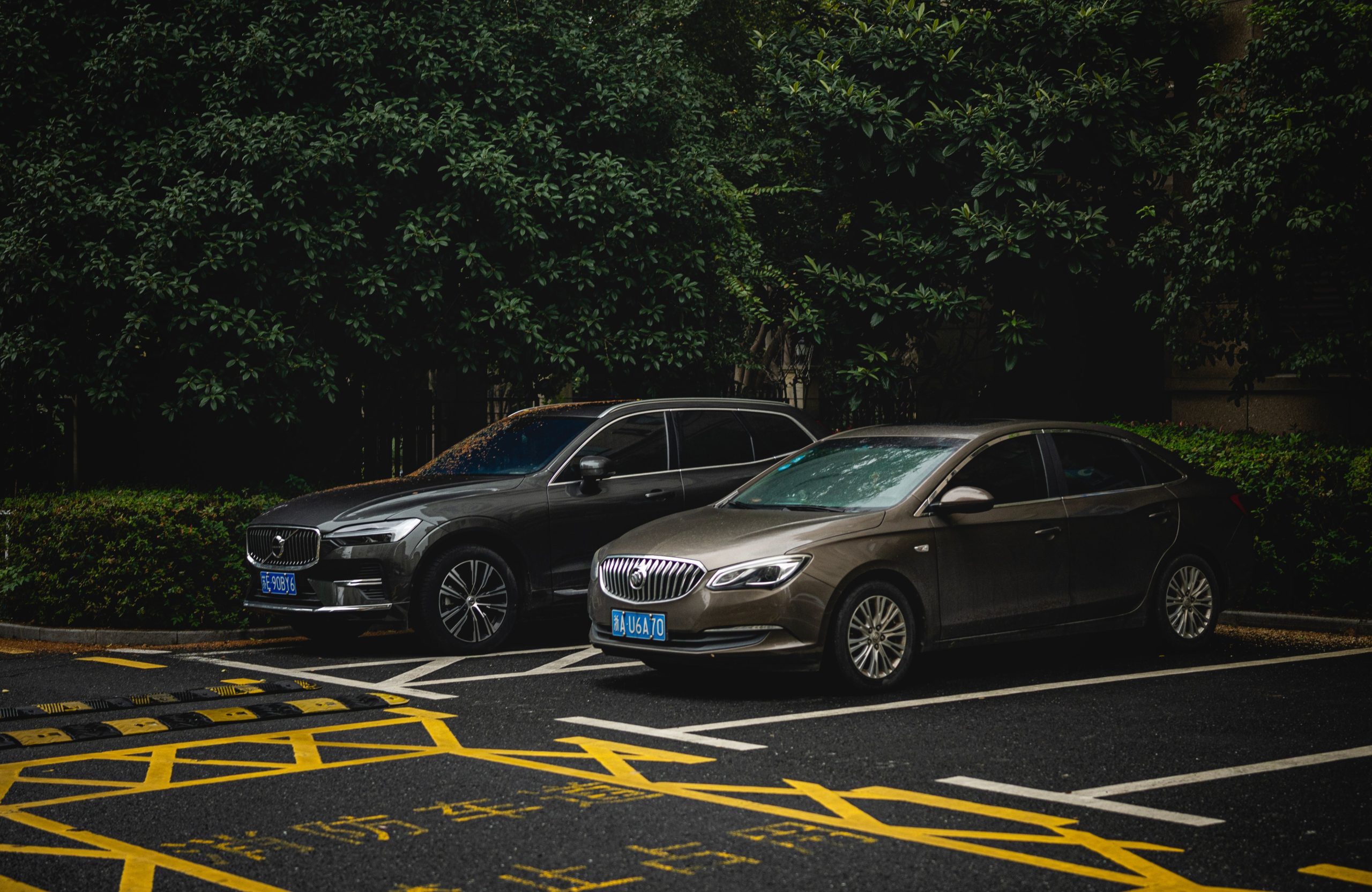 Two cars, an SUV and a sedan, parked side by side in a shady urban lot. Deux voitures, un SUV et une berline, garées côte à côte dans un parking urbain ombragé.